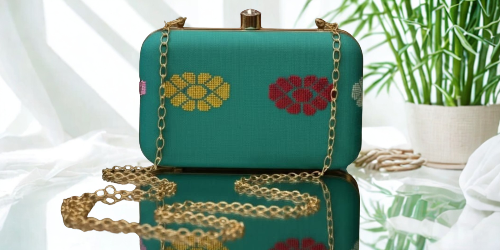 Teal clutch bag with floral embroidery on a reflective surface with a plant in the background