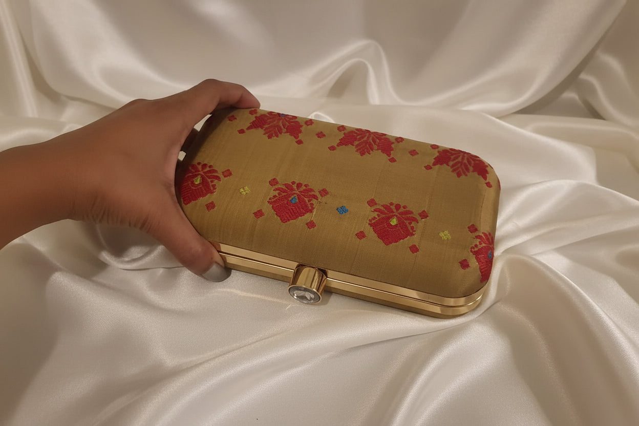 Handwoven Assam silk clutch bag in antique gold colour with red traditional Assamese motifs and subtle multicolour detailing, featuring a metal clasp and structured frame