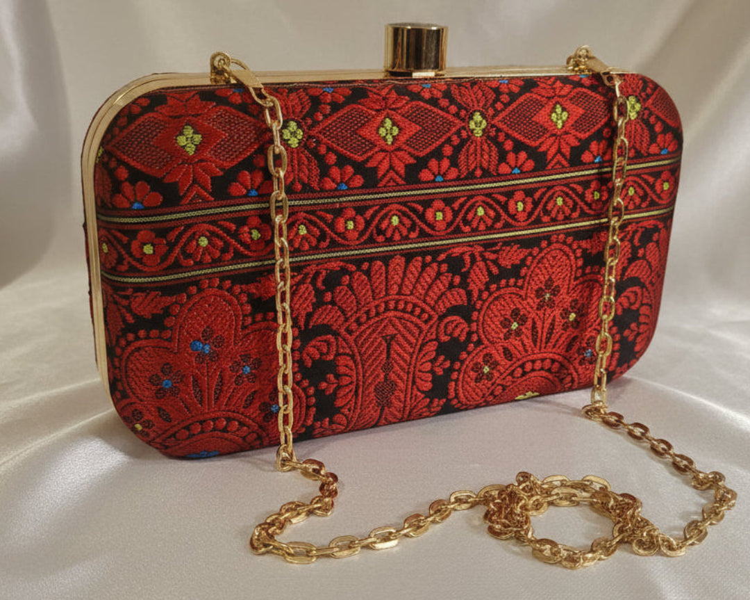 Handwoven Assam silk clutch bag in black and red colour with traditional Assamese motifs, gold metal frame and detachable chain, styled on a tabletop background