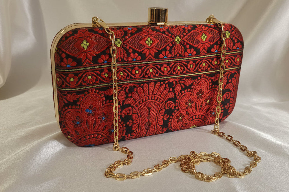 Handwoven Assam silk clutch bag in black and red colour with traditional Assamese motifs, gold metal frame and detachable chain, styled on a tabletop background