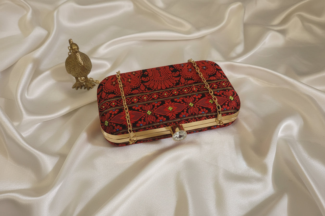 Handwoven Assam silk clutch bag in black and red colour with traditional Assamese motifs, gold metal frame and detachable chain, styled on a tabletop background